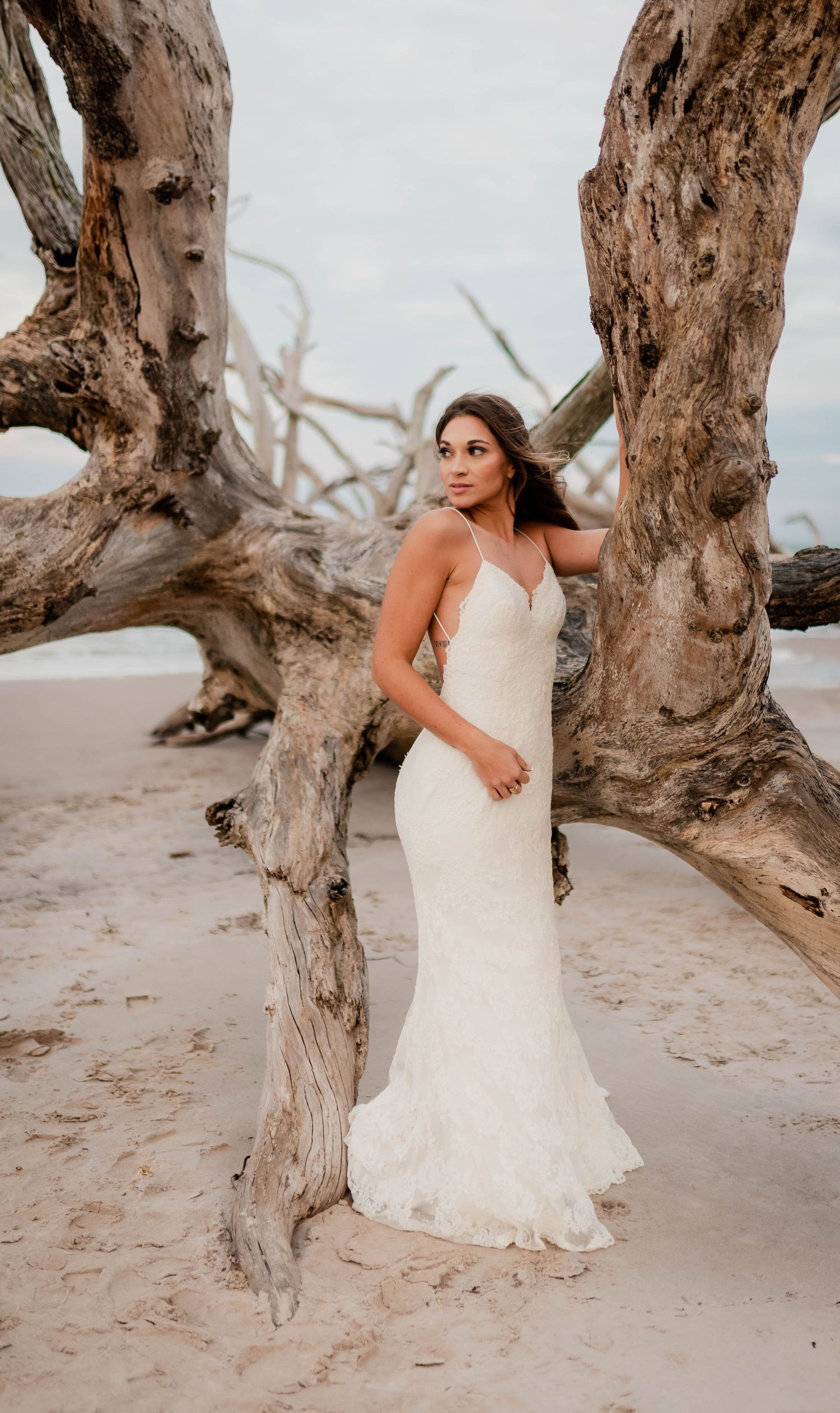 Bride in white lace mermaid gown poses among driftwood on sandy beach, showcasing elegant spaghetti strap wedding dress.
