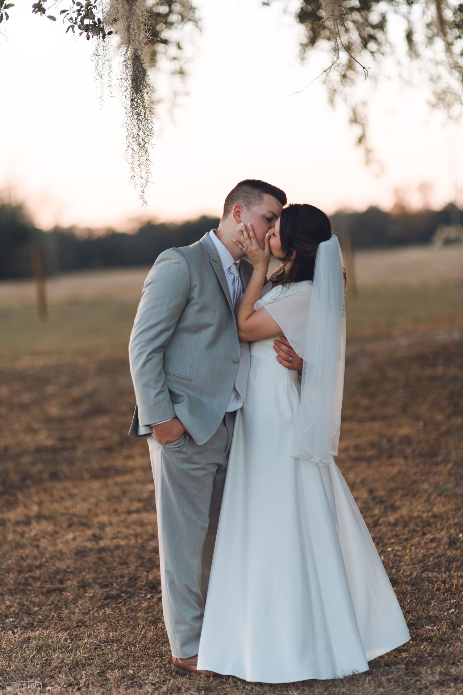 Bride and groom sharing a romantic kiss at sunset in an outdoor field, bride in white gown with veil, groom in gray suit.