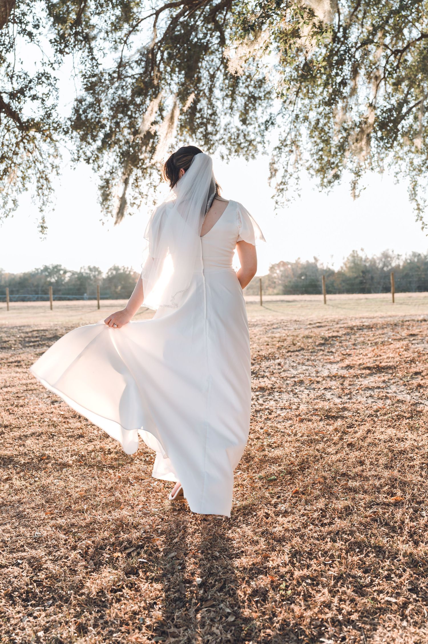 Bride in white wedding dress and veil twirling outdoors under oak tree in sunlit field.