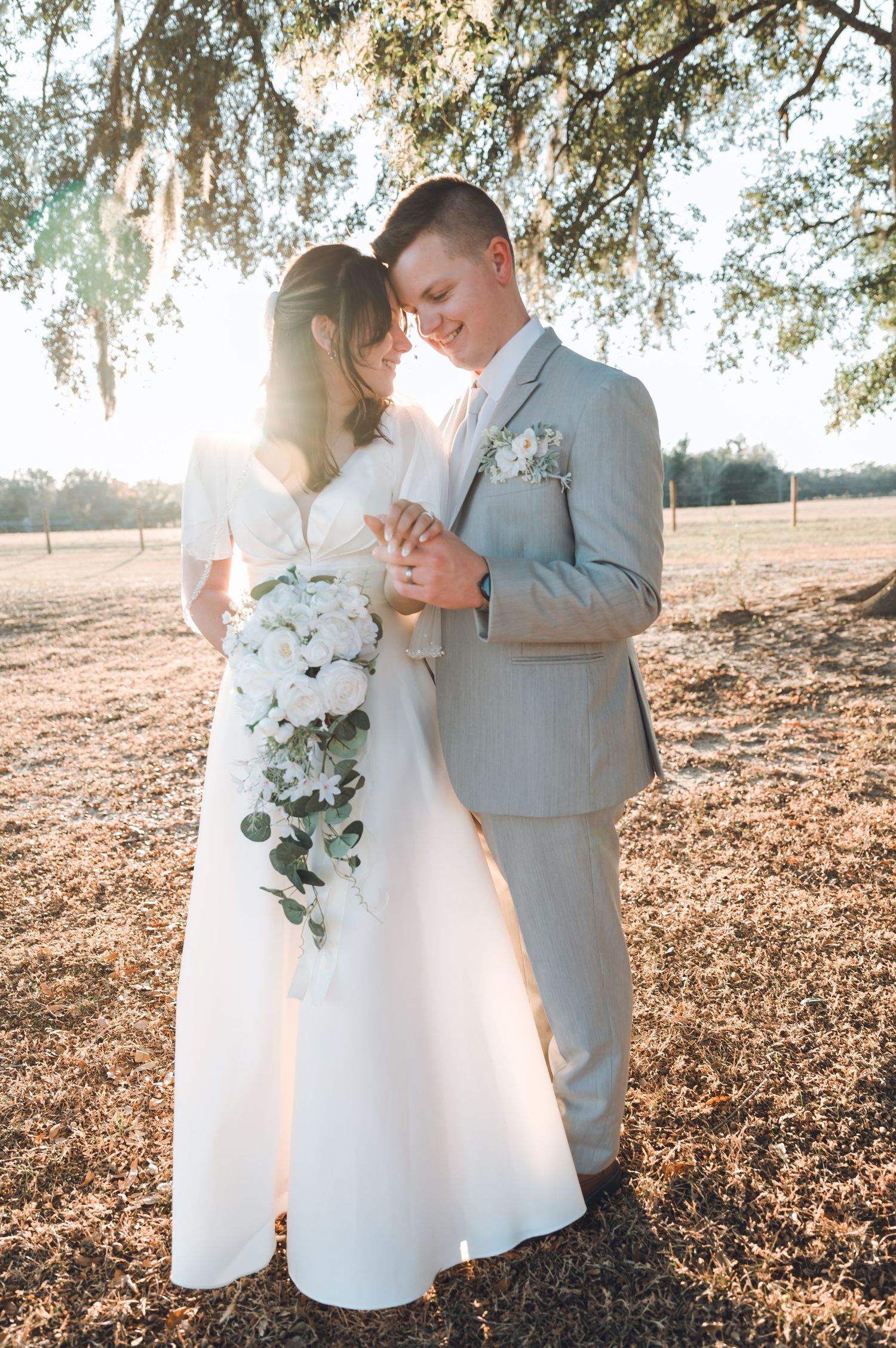 Bride and groom share a tender moment under oak tree, golden sunlight glowing behind them, white floral bouquet.