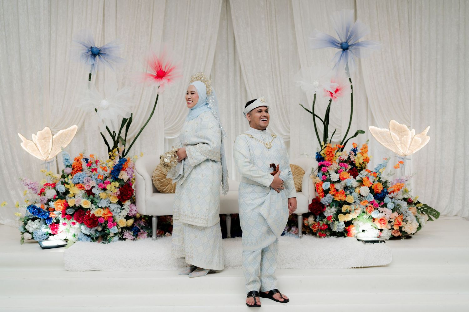 Muslim couple in white attire at a colorful floral wedding ceremony backdrop with decorative flowers.