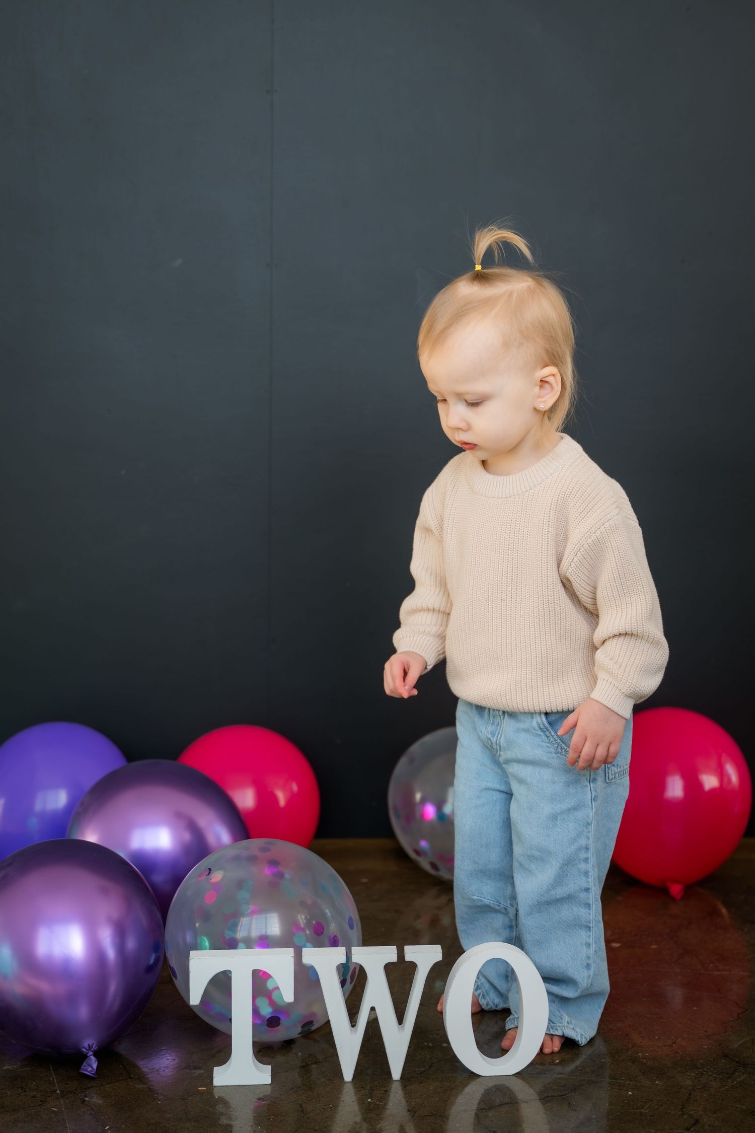 Toddler in beige sweater and jeans stands by colorful balloons and TWO sign for second birthday photo.