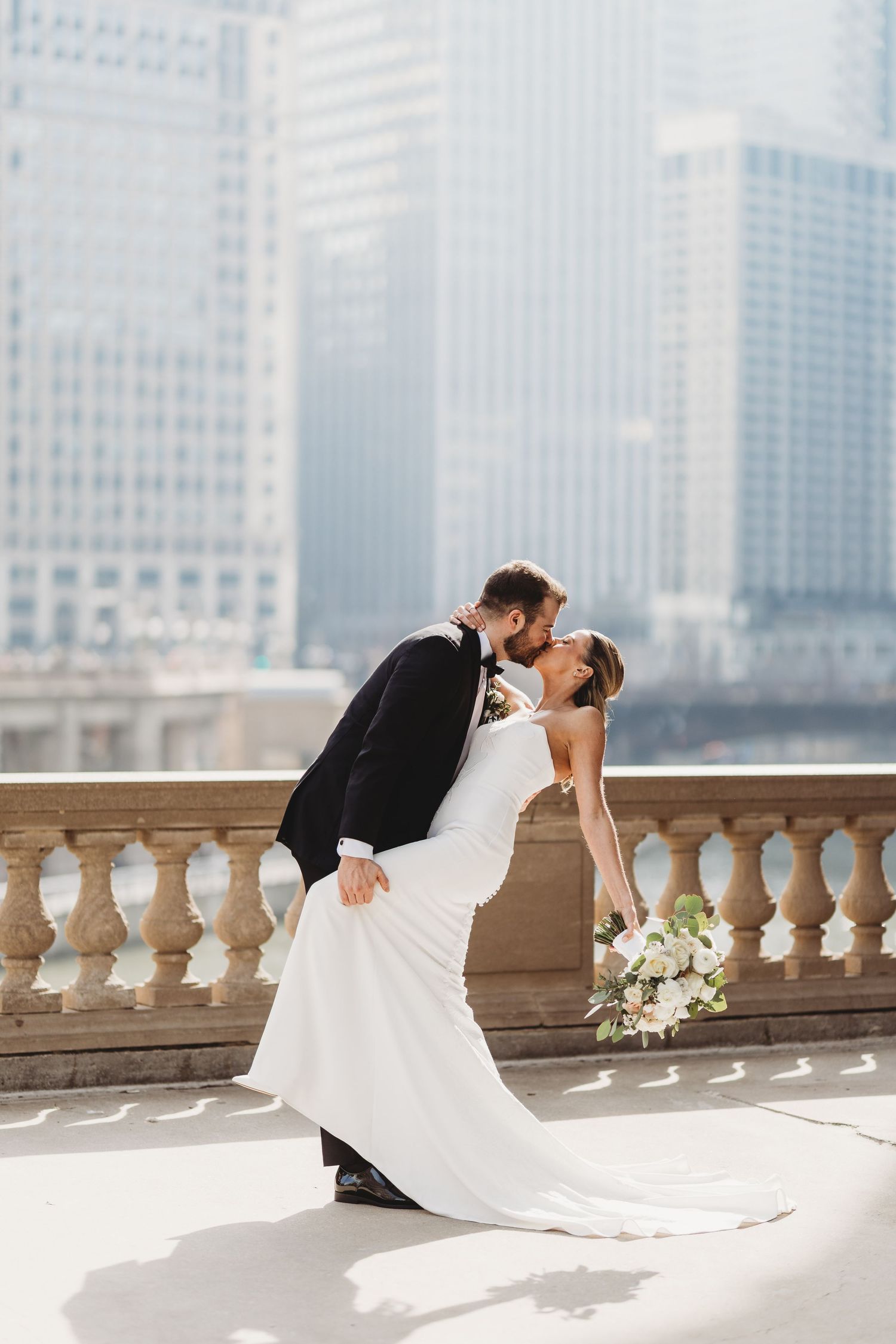 Bride and groom share a romantic dip kiss on a city terrace, bride holding white bouquet in elegant wedding attire.