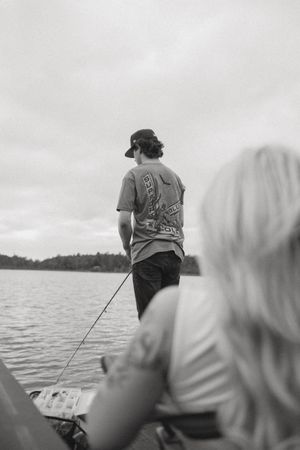 A man fishing from a boat on a calm lake while a blonde woman watches in this black and white photo.