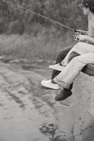 Black and white photo of two people sitting on a rock fishing together with a rod over a calm body of water.