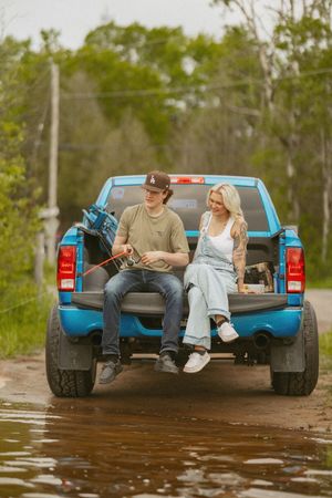 Young couple sitting on tailgate of blue pickup truck on a muddy rural road surrounded by green trees.