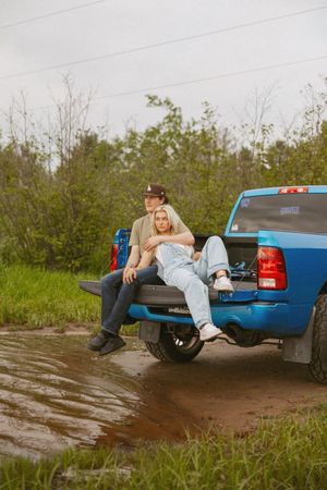 Couple sitting on tailgate of blue pickup truck on muddy rural road surrounded by green trees.