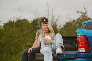 Couple sitting together on the tailgate of a blue pickup truck surrounded by lush green trees.