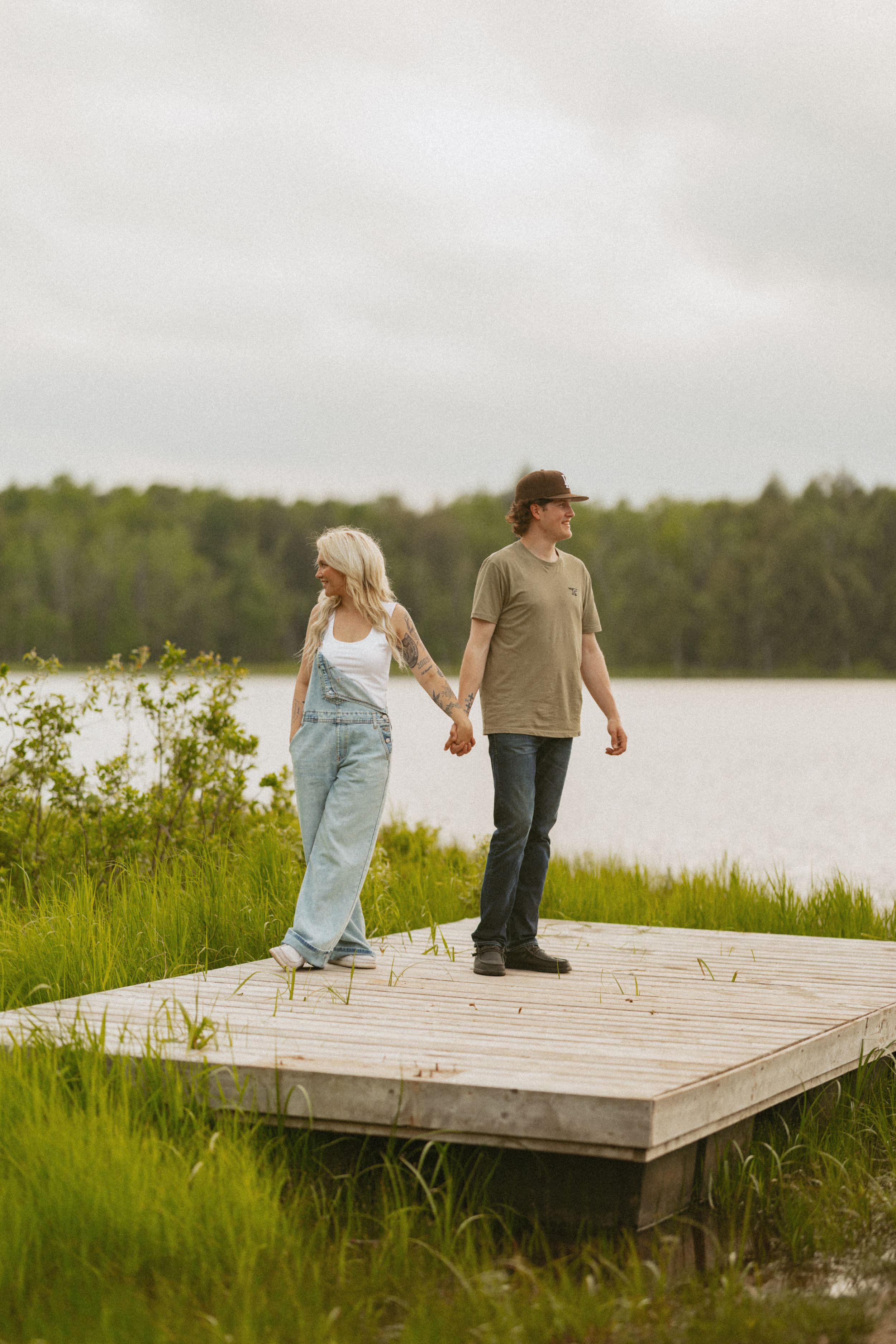 Young couple holding hands on a wooden dock by a calm lake surrounded by lush green landscape.