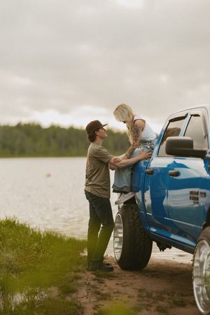 A couple sharing a romantic moment by a blue pickup truck near a serene lake on a cloudy day.