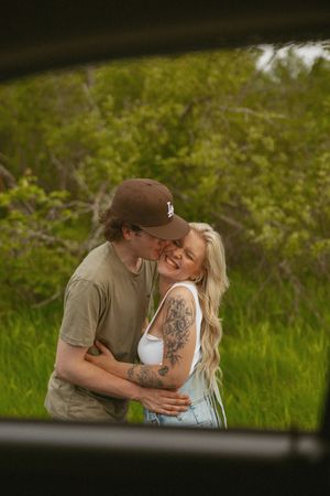 A happy couple embracing and kissing outdoors surrounded by lush green trees and foliage.