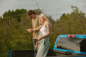 Couple fishing together outdoors near a blue truck, surrounded by lush green trees on a sunny day.