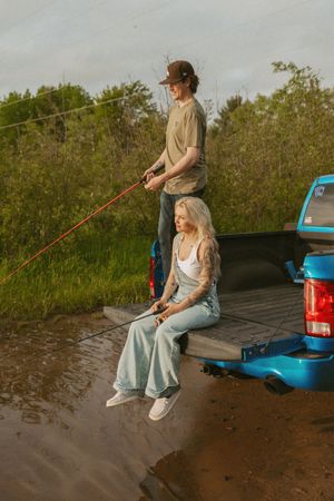 Couple fishing from the back of a blue pickup truck parked by a muddy river at sunset.