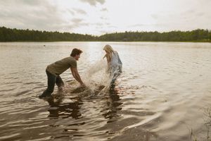 Playful couple splashing water at each other while standing in a serene lake with forest backdrop.