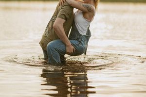 Romantic couple embracing and kissing while standing in shallow water, wearing jeans and casual clothes.