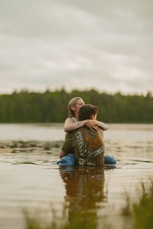 Romantic couple embracing in a lake, woman with tattoos hugging man in wet clothes under cloudy sky.
