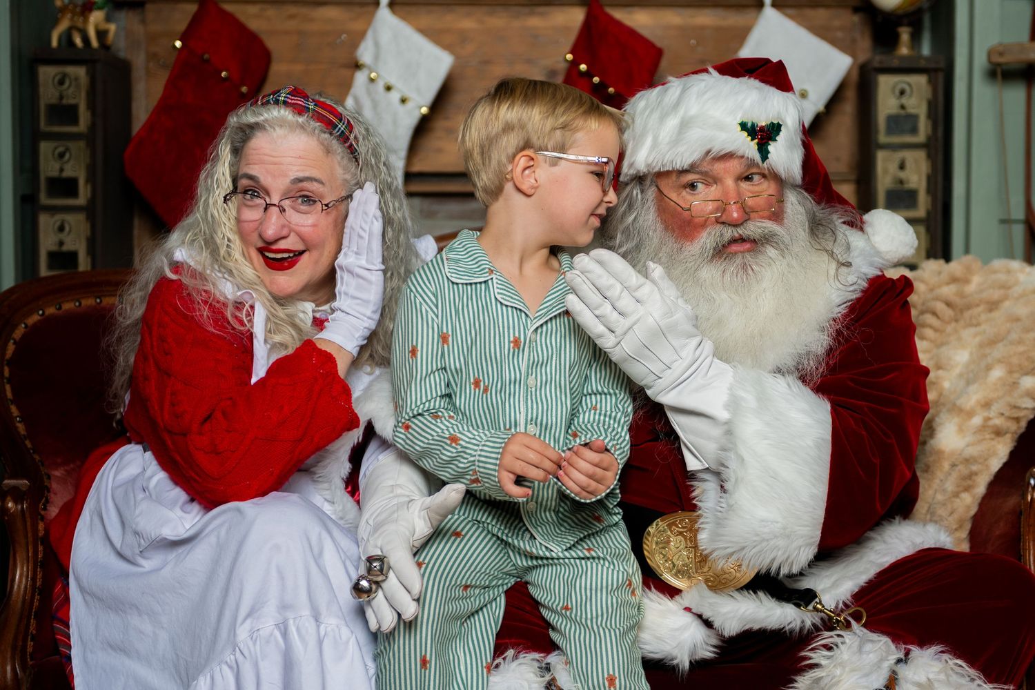 Child in pajamas smiling with Santa Claus and Mrs. Claus in festive holiday setting with stockings.