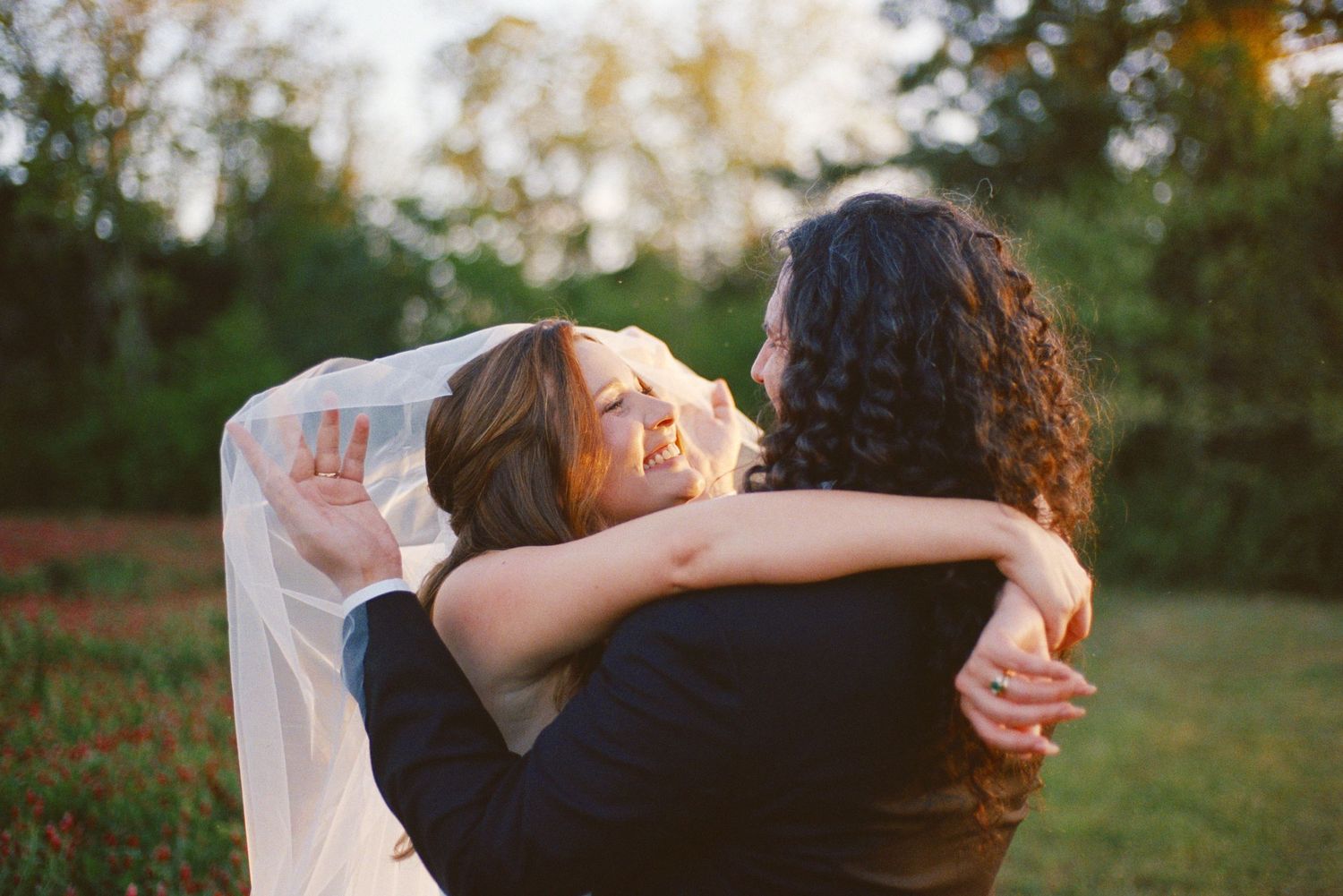 Bride in veil embracing someone in a warm outdoor hug, smiling joyfully at a wedding celebration.