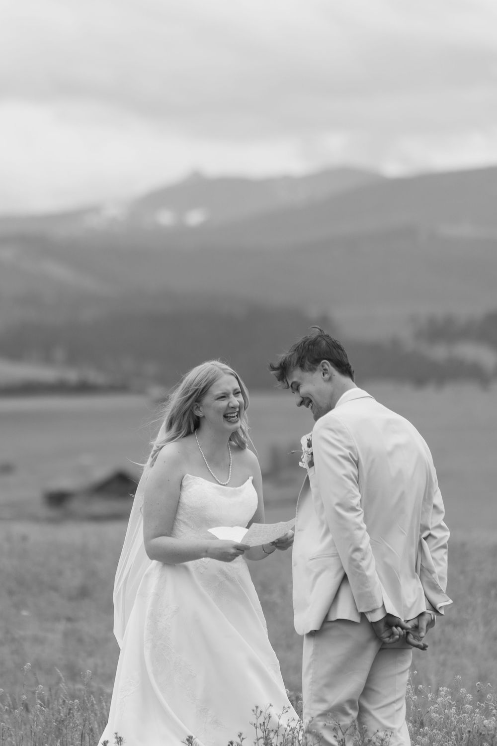 Black and white photo of a laughing bride and groom in a field with mountain backdrop on their wedding day.