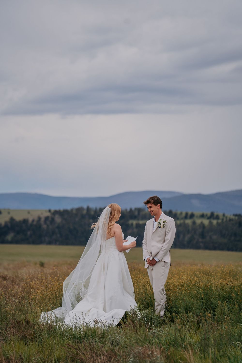 Bride and groom exchange vows in a scenic mountain meadow, bride in white gown with veil, groom in beige suit.