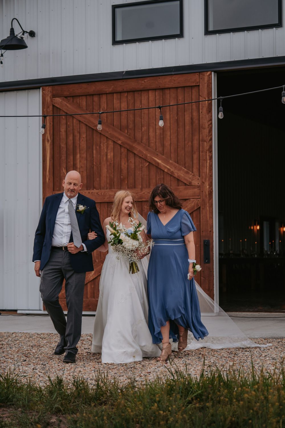 Bride walks with her parents out of a white barn venue, holding a bouquet on her wedding day.
