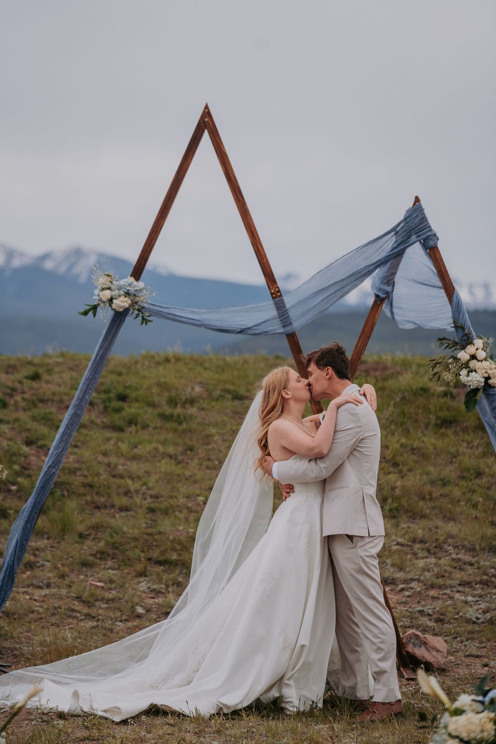 Bride and groom share first kiss under a triangular wooden arch with blue draping and mountain backdrop.