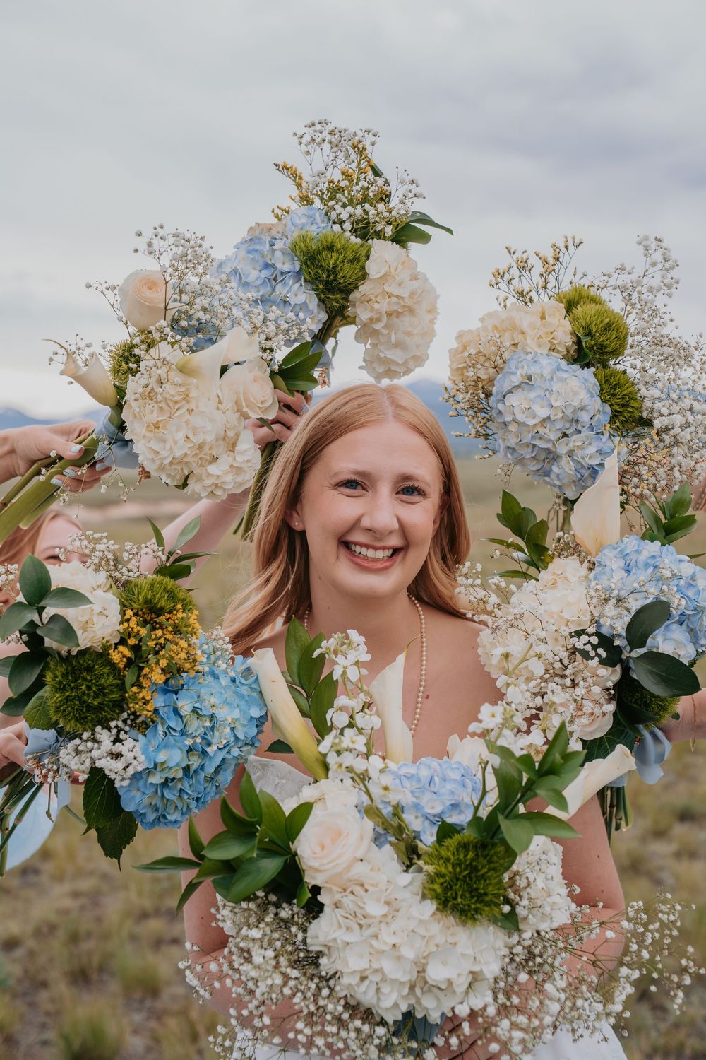 Smiling bride surrounded by blue and white hydrangea bouquets held by bridesmaids outdoors.