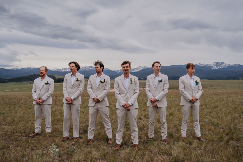 Six groomsmen in tan suits stand in a mountain meadow under cloudy skies for a rustic outdoor wedding.