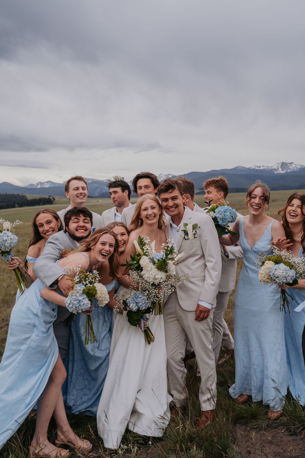 Wedding party in blue dresses and white suits celebrate outdoors with mountain backdrop and floral bouquets.