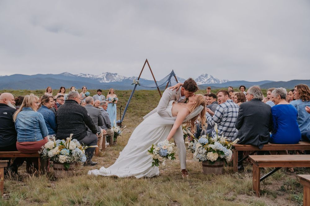 Bride and groom share first kiss at outdoor mountain wedding ceremony with snow-capped peaks backdrop.