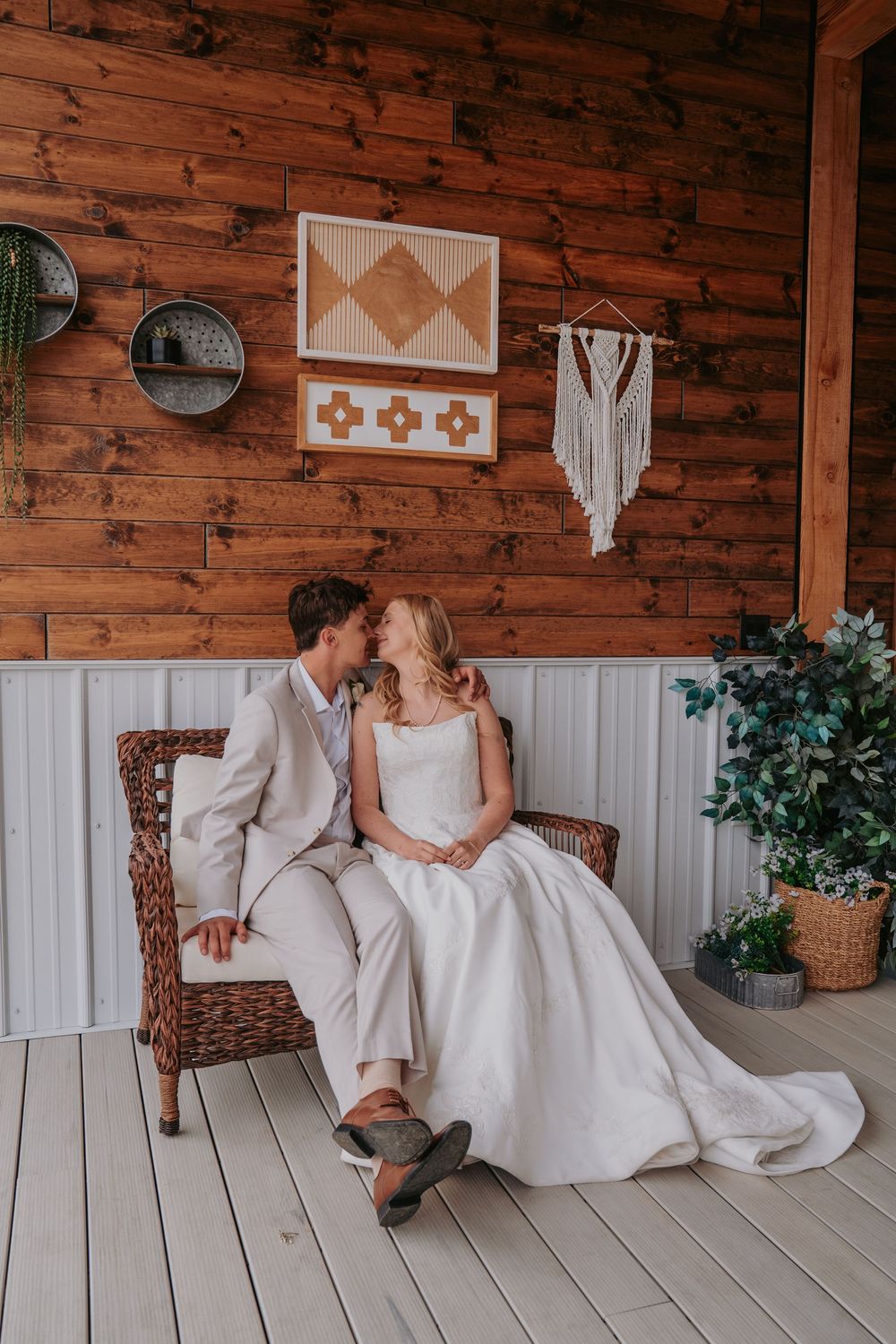 Bride and groom share a romantic moment on a rustic wooden porch, seated in a wicker chair with boho wall decor.