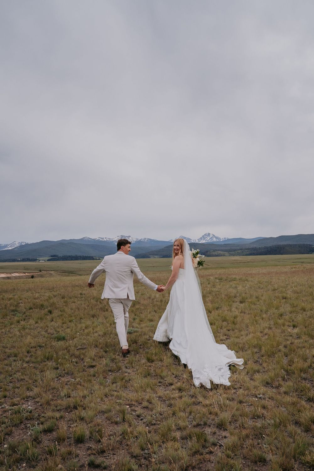 Bride and groom holding hands walking through a green meadow with snow-capped mountains in the background on their wedding day.