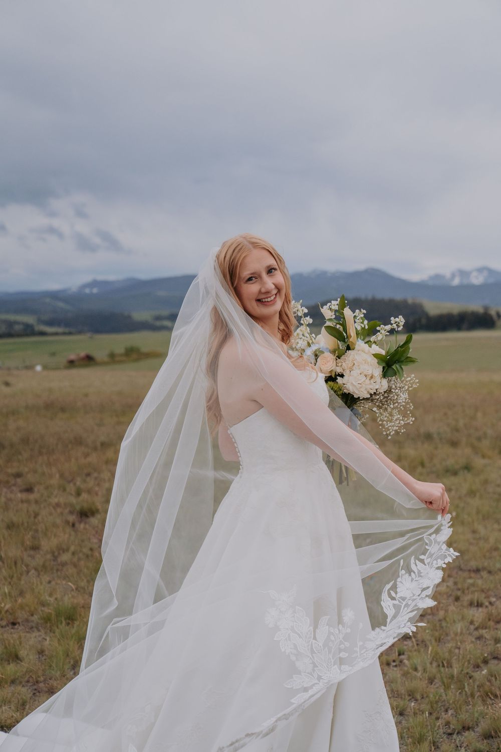 Smiling bride in a white gown and lace veil holds a bouquet in an open field with mountain views behind her.