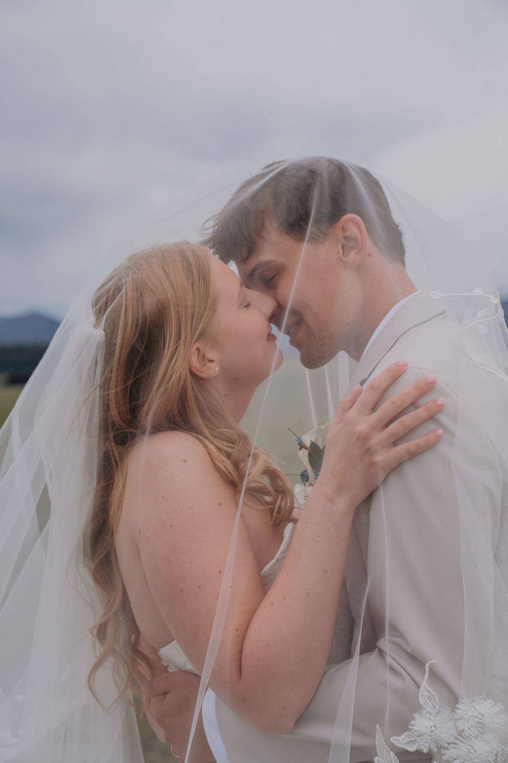 Bride and groom share an intimate moment under a flowing white veil outdoors on their wedding day.
