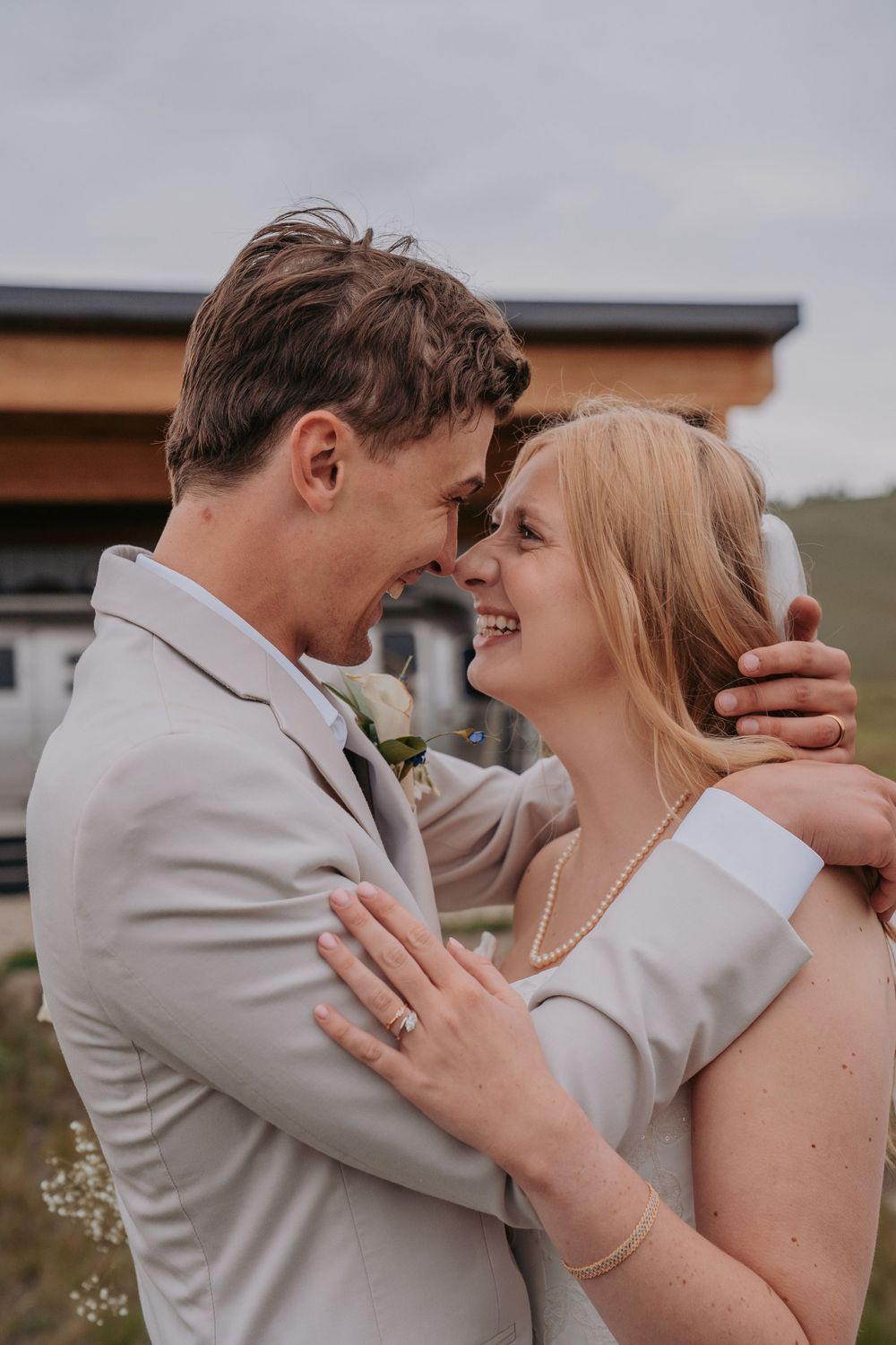 A smiling bride and groom in wedding attire embrace outdoors, touching foreheads in a romantic moment.