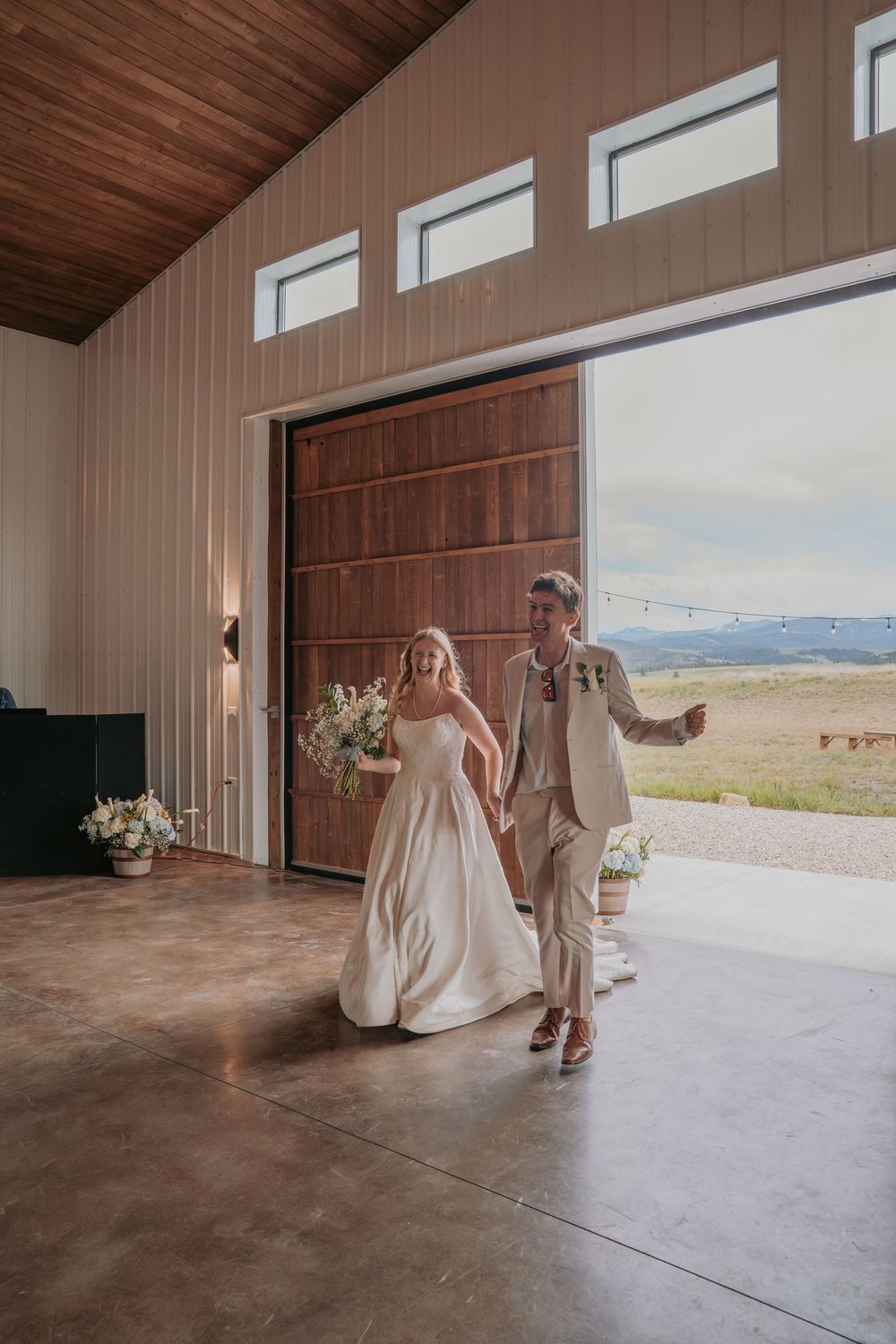 Bride and groom walk hand in hand through a rustic barn venue with mountain views on their wedding day.