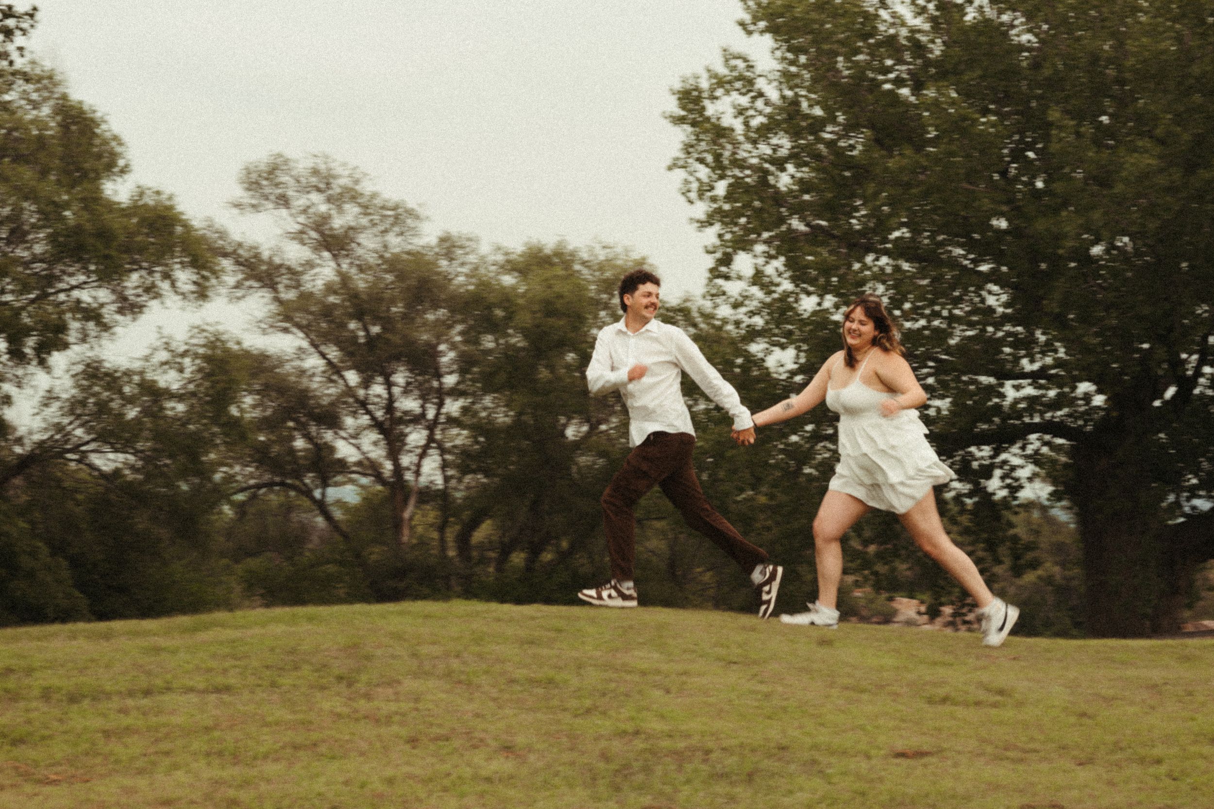 Whimsical outdoor couples session featuring a young couple in white running with motion blur surrounded by soft greenery and natural light.