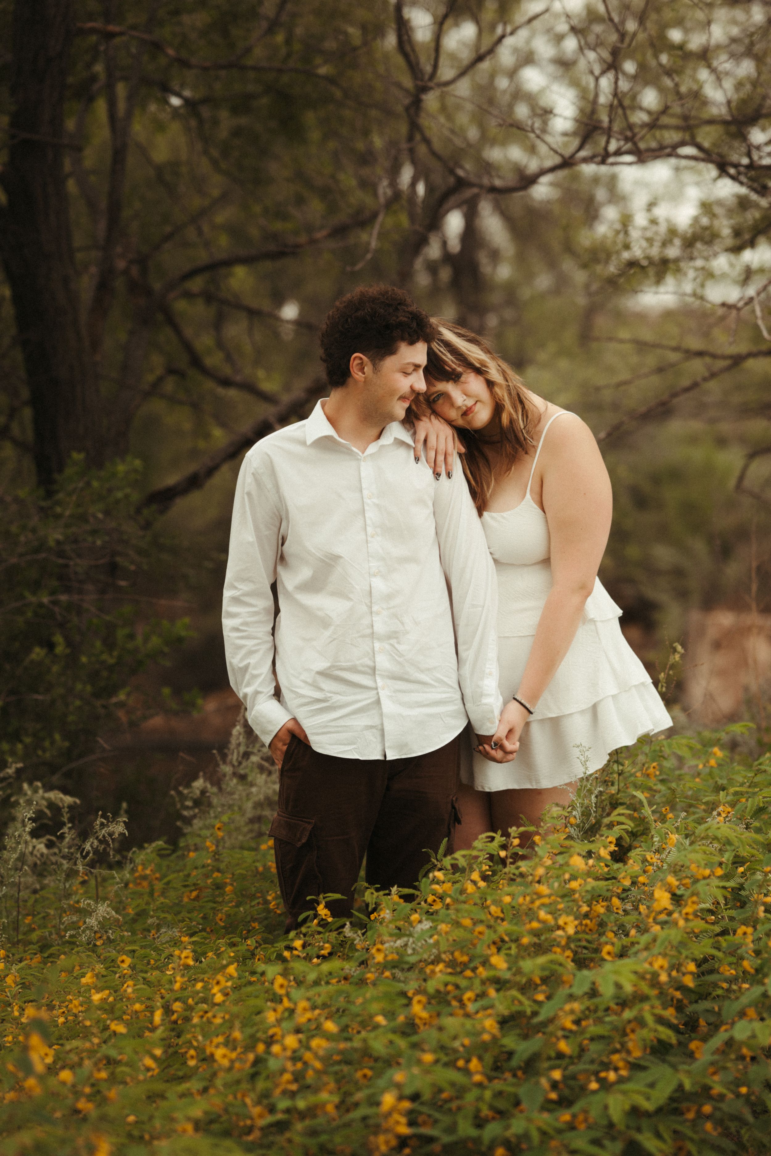 Romantic couples portrait with a woman in a white dress and a man in a white shirt in a lush green setting.