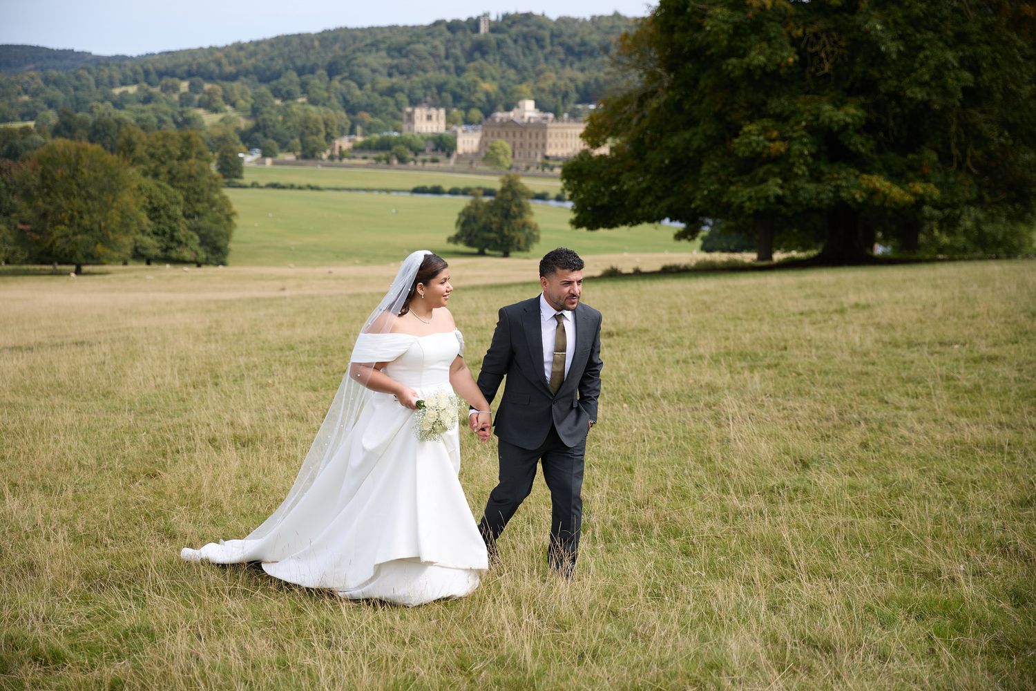 Bride and groom walk hand in hand through a golden field with a grand estate and rolling hills behind them.