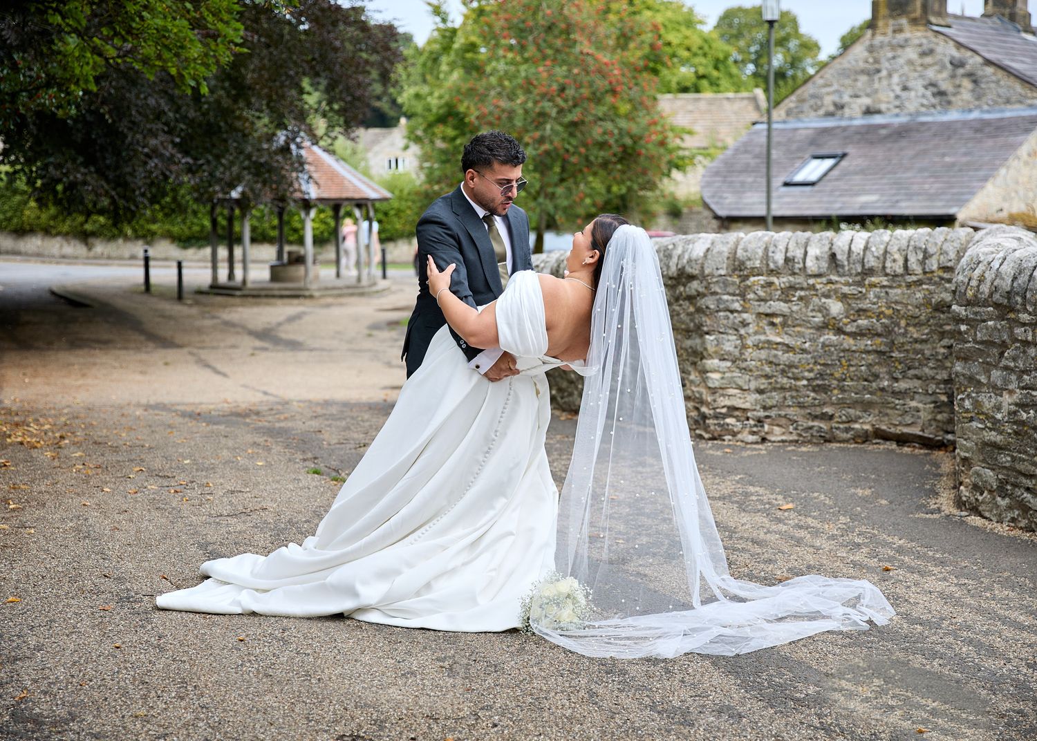 Bride and groom share a romantic dip kiss in white wedding dress and veil outside a stone building.