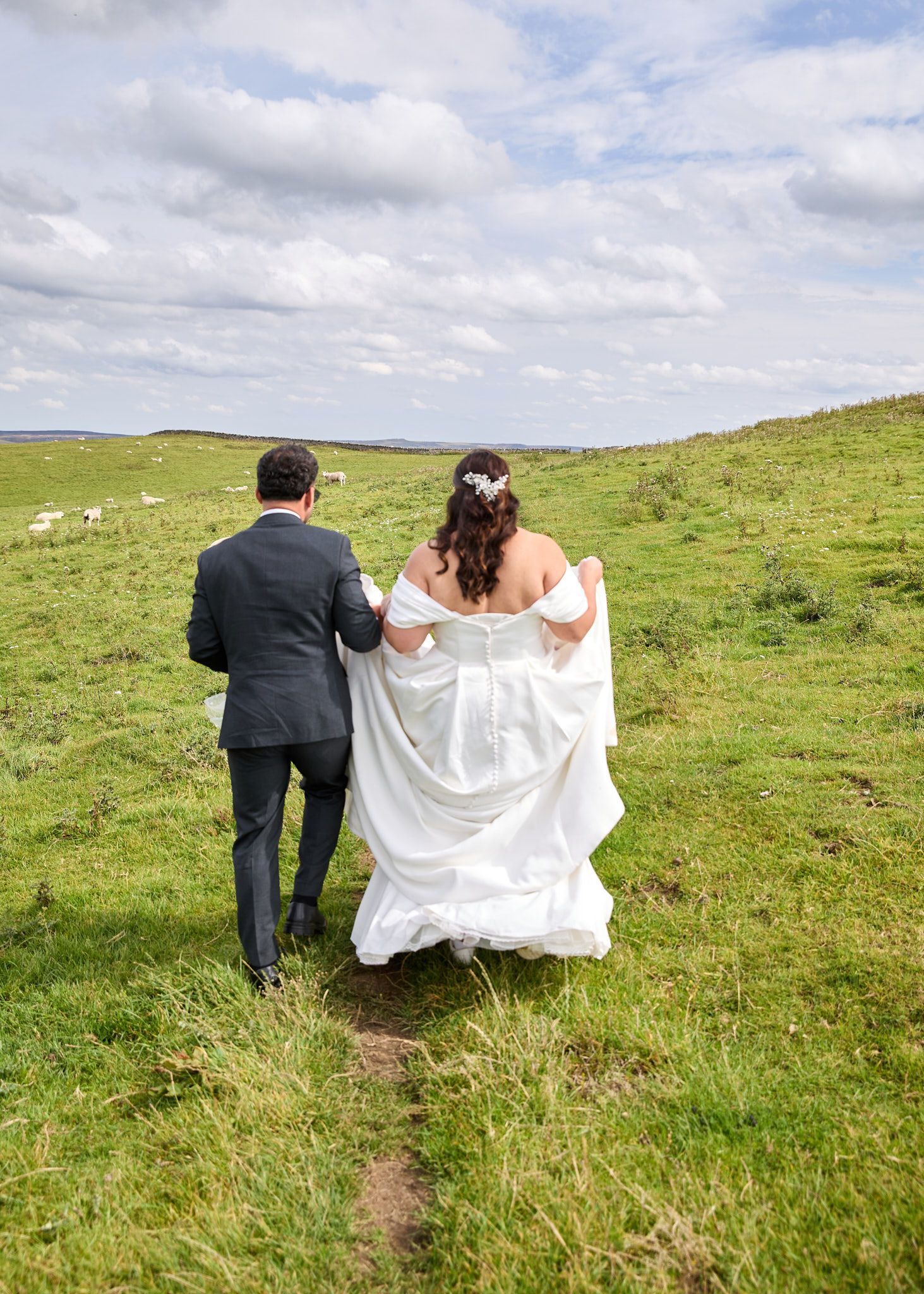 Bride and groom walking hand in hand across green countryside fields on their wedding day.