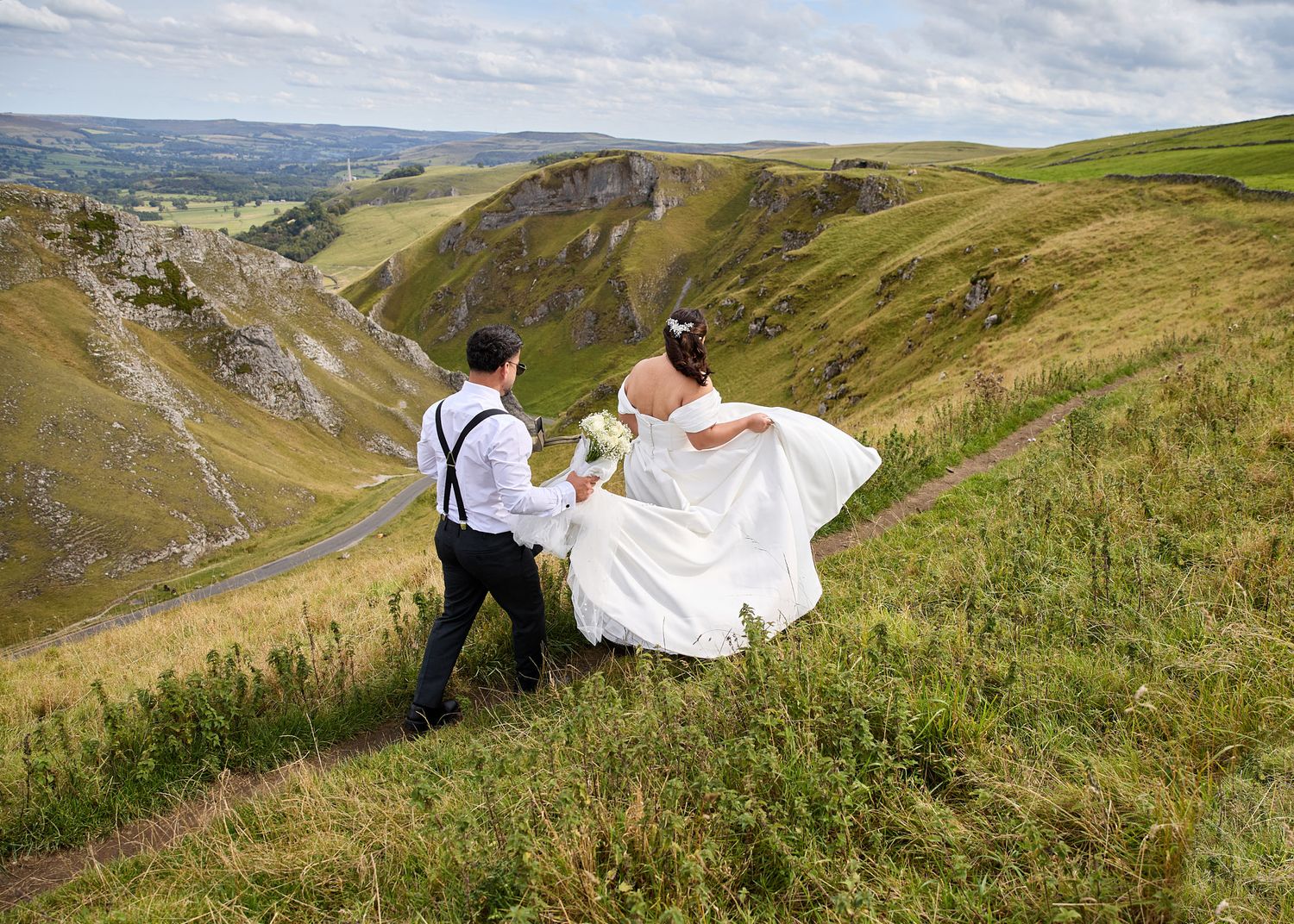 Bride and groom walk hand-in-hand along a lush green hillside with a stunning valley landscape backdrop.
