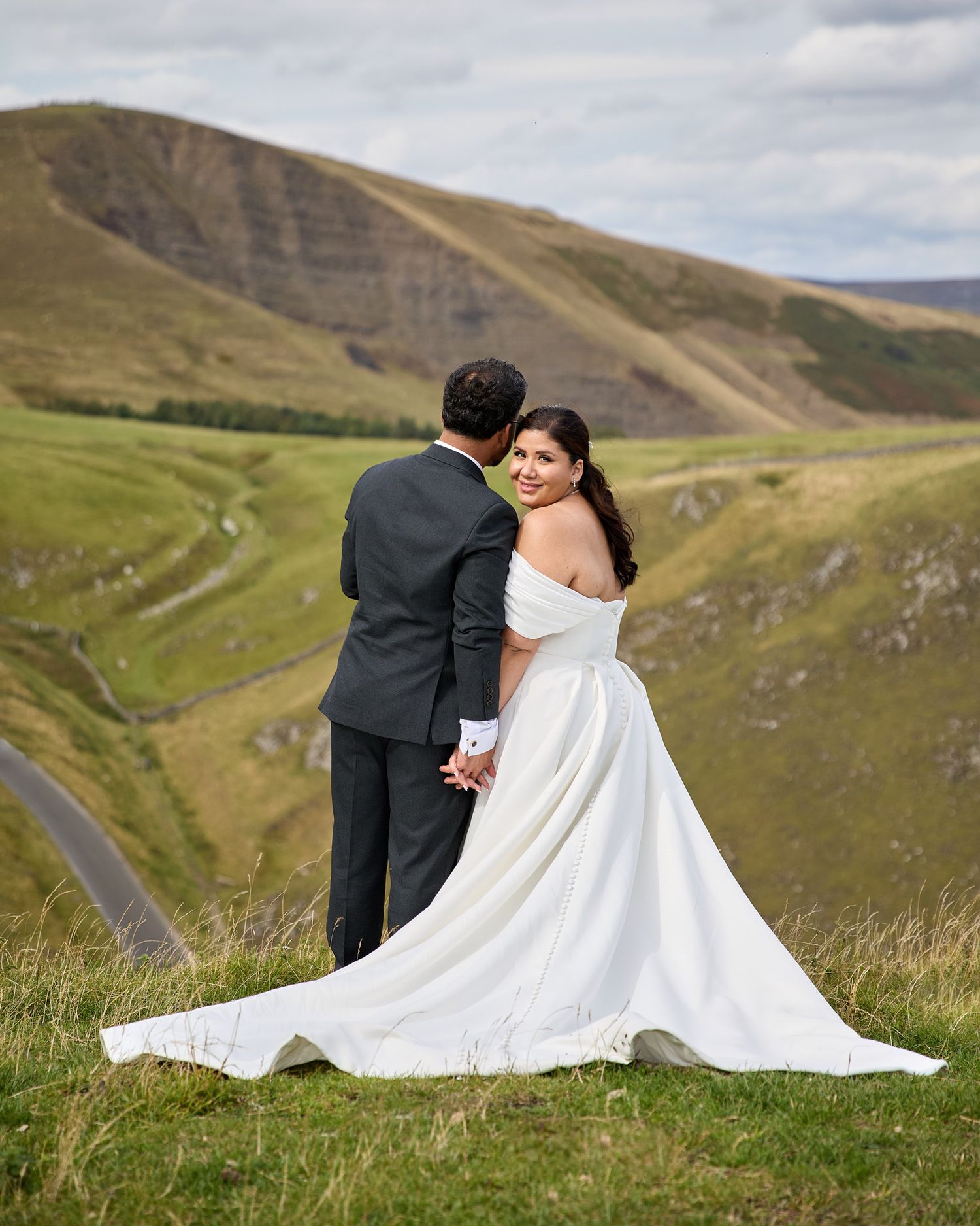 Bride in off-shoulder white gown and groom in dark suit hold hands on a scenic green hillside.