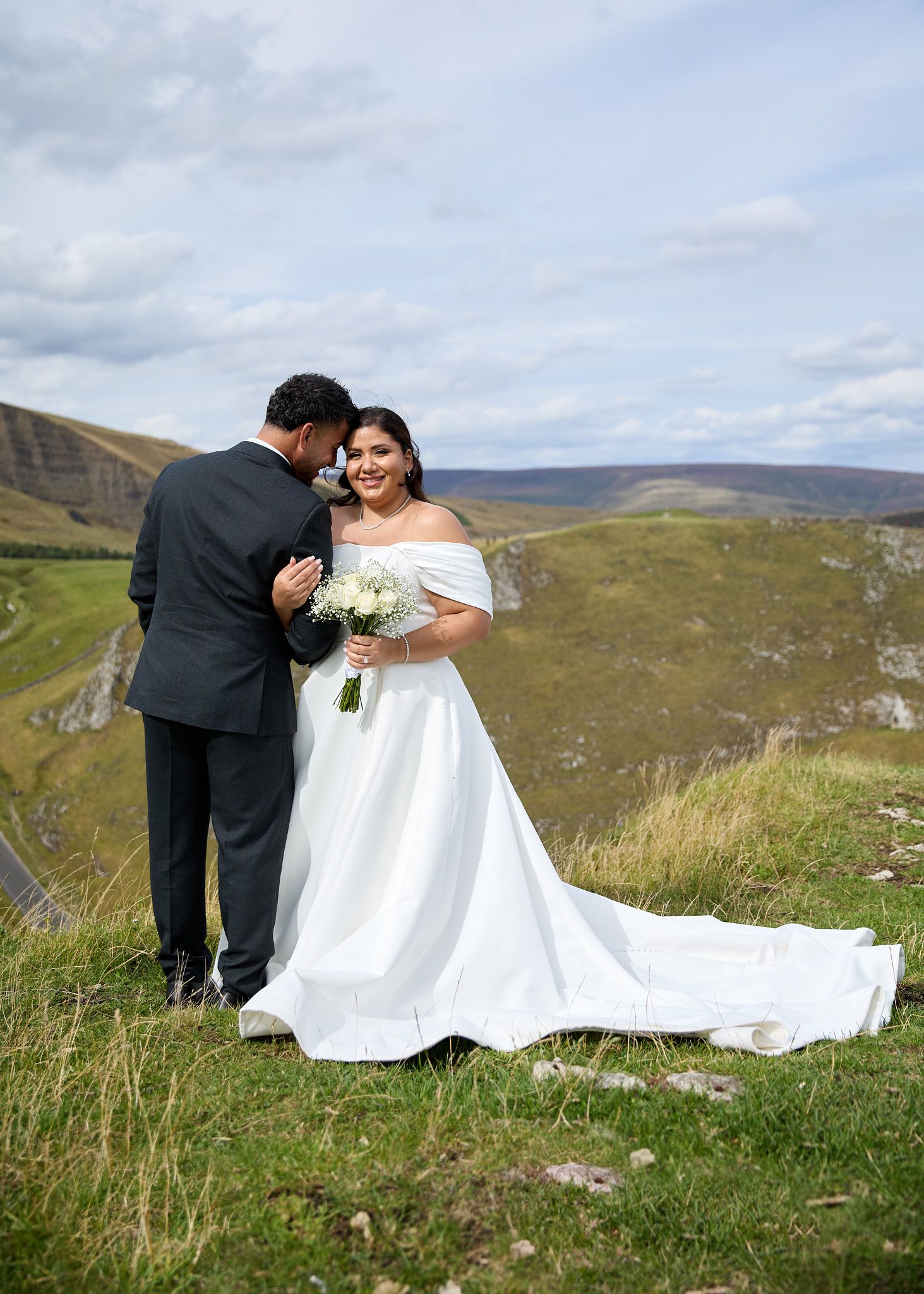 Bride in off-shoulder white gown holds bouquet as groom kisses her cheek on scenic green hillside.