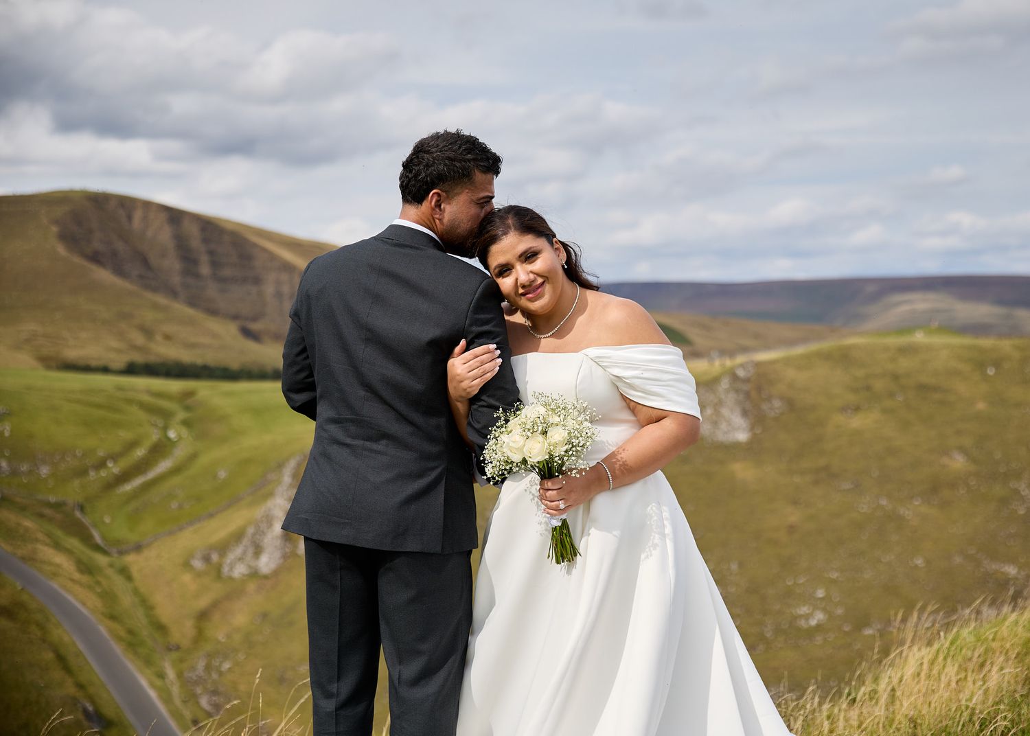 Bride and groom embrace on a scenic hilltop in wedding attire, bride holding white bouquet with rolling green hills behind.