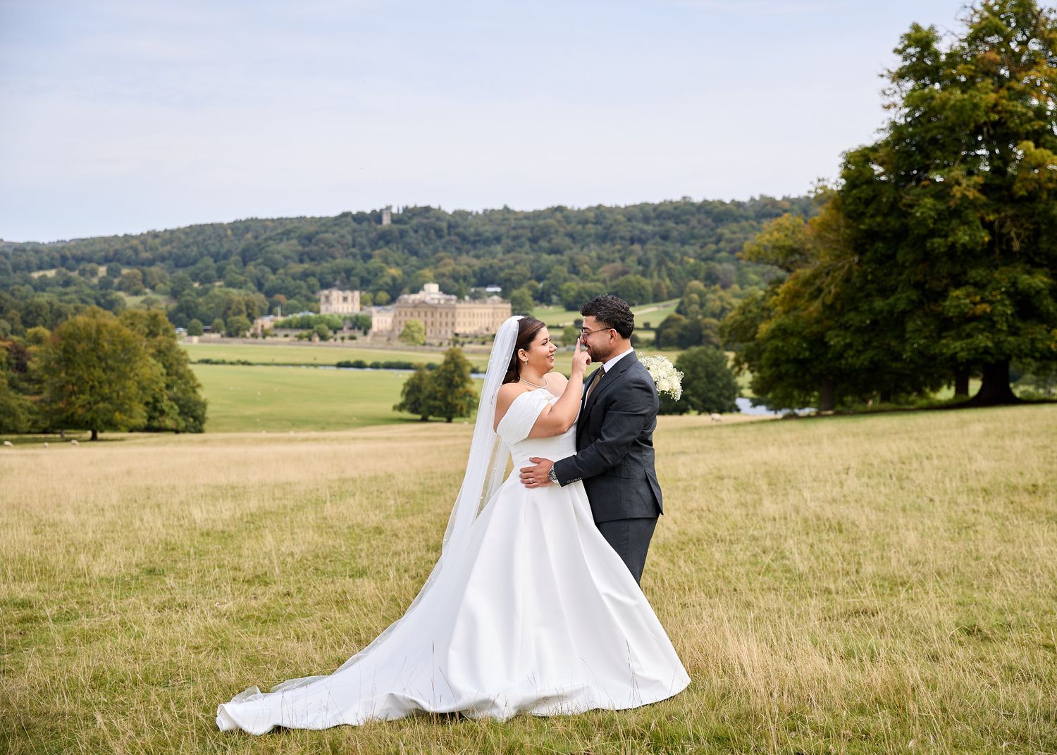 Bride and groom embrace romantically in a sunlit meadow with a stately manor and lush hillside in the background.
