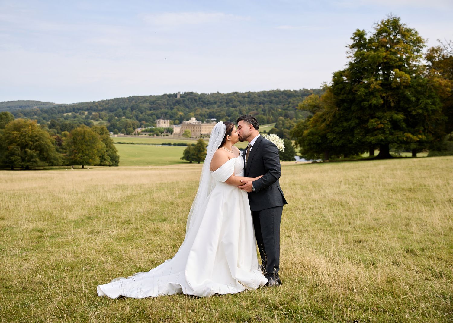 Bride and groom kissing in a scenic countryside field, with rolling hills and trees in the background on their wedding day.