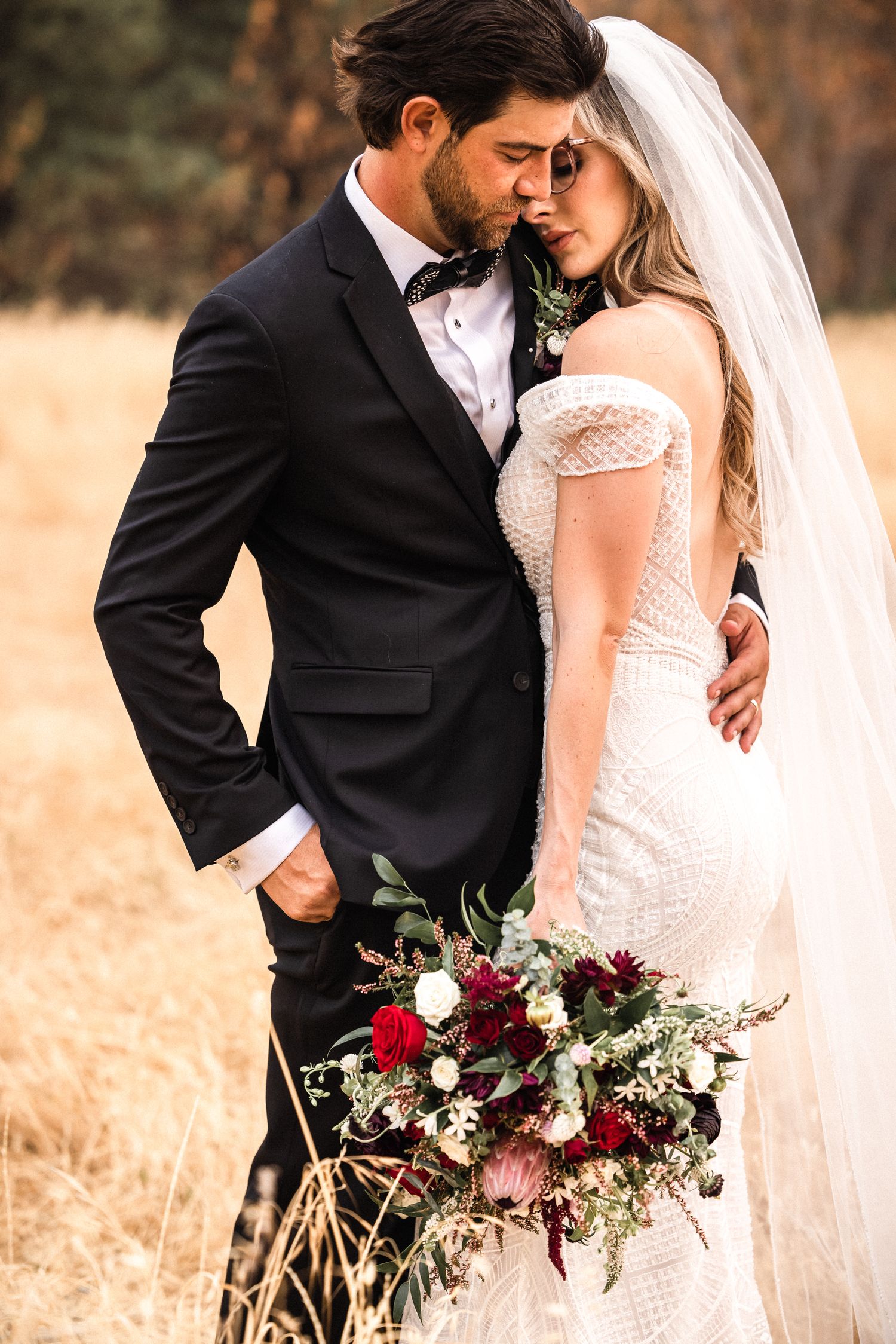 A bride and groom share an intimate moment outdoors, she holds a lush red and white bouquet while wearing a lace gown.