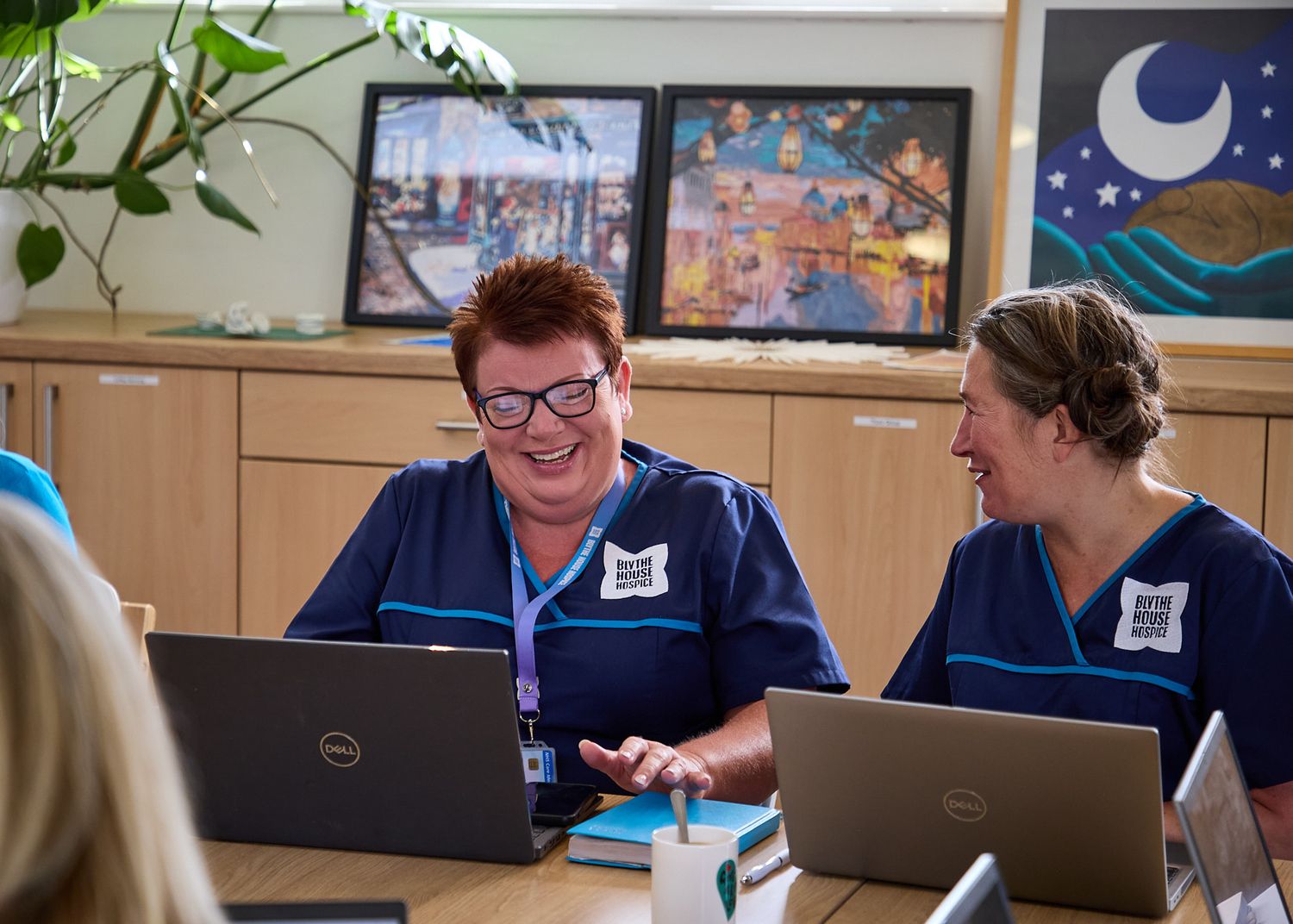 Two nurses in blue scrubs smile while working on laptops together in a bright healthcare office environment.
