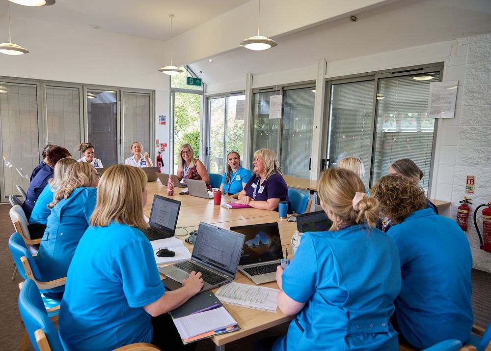 Healthcare professionals in blue scrubs collaborate around tables with laptops during a medical team meeting in a modern office.